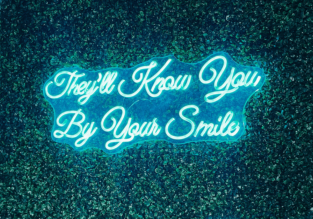 They'll Know You By Your Smile cursive led sign on a faux greenery background
