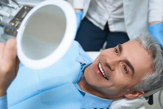 Smiling man in a dental chair holding a mirror, wearing a blue bib, with a dentist in the background, Showing satisfaction after a successful gum disease treatment