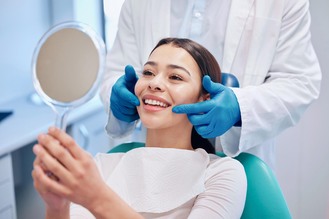 Patient in dental chair smiling into handheld mirror while dental professional in blue gloves gently checks face in bright clinical setting.